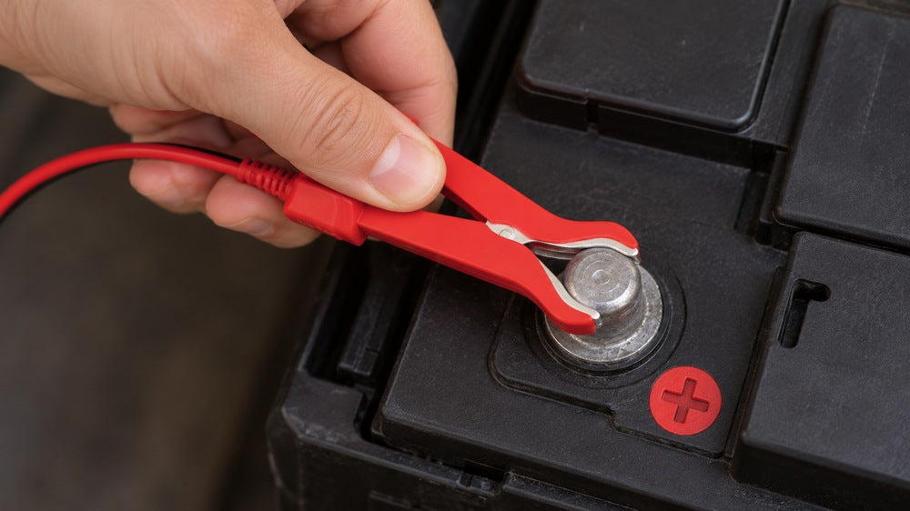 A hand connects the red clamp to a car battery positive terminal